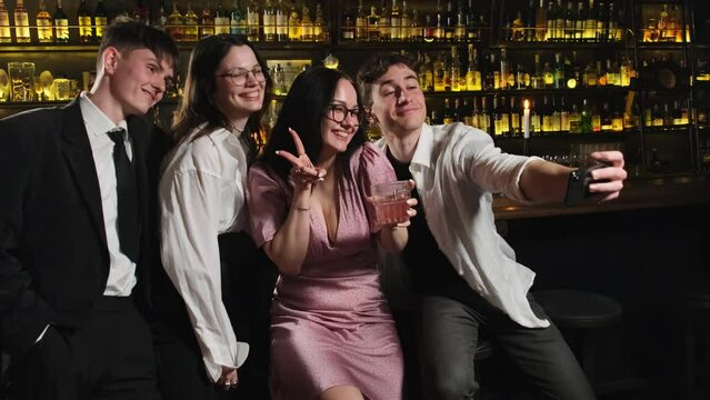 Positive woman in pink satin dress makes kiss lips for joint photo. Young people have fun at bar counter in contemporary pub