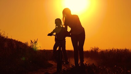 Child dream of riding bike. Mother teaches her child to keep balance while sitting on bicycle. Mom teaches her child, little daughter to ride bike, sunset. Family life, mom, baby, parental support.