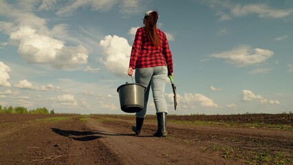 Agricultural industry. Farmer woman working in the field in spring. Farm business. Farmer woman walking along dirt road plowed field with shovel, bucket. Gardener walking with bucket, shovel in field