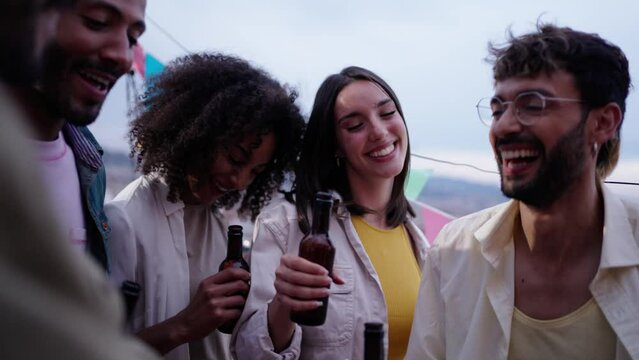 Cheerful multiracial young people toast with beer bottles celebrating dancing and laughing on summer outdoors. Excited group happy millennial friends together enjoying sunset drinks at rooftop party 