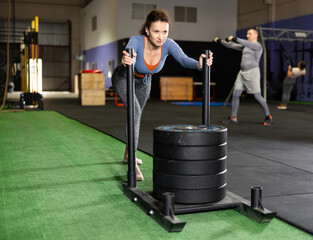Focused middle-aged woman in sportswear pushing heavyweight plates on prowler slider in gym