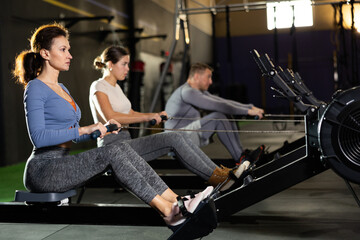 Concentrated sporty adult woman working out on rowing machine during total-body workout in gym....
