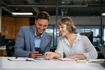 Two caucasian business people looking down and smiling while using smart phone in office
