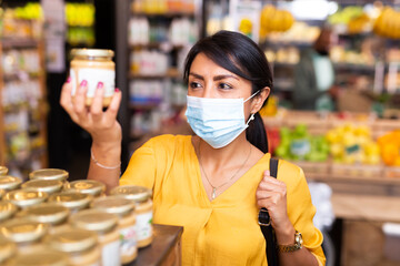 Woman in protective mask reads description of canned food at a grocery supermarket
