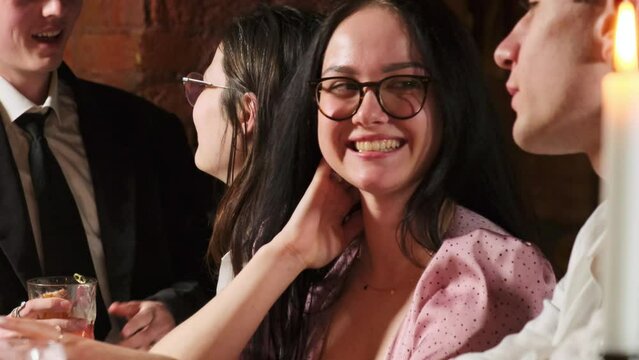 Woman with nice smile flirts with guy in romantic atmosphere. Young people sit at bar counter with pretty couple in background