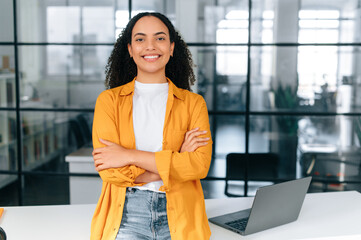 Photo of a gorgeous positive confident brazilian or hispanic curly haired young woman in a casual stylish clothes, stand in the office near workdesk with arms crossed, looks at camera, smiling happily