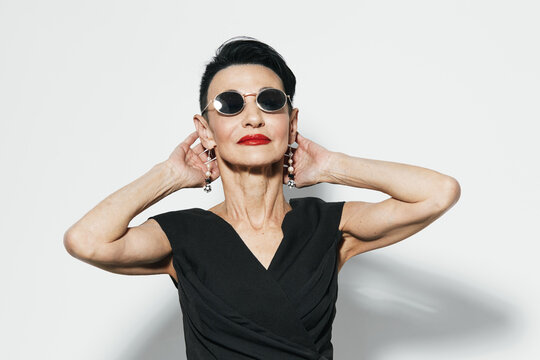 Fashionable woman in sunglasses and black dress posing with hands on head against white wall, elegant and stylish look