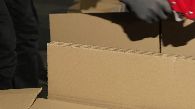 Man worker uses parcel tape gun to secure empty cardboard boxes. Factory employee in gloves prepares packaging for ready products