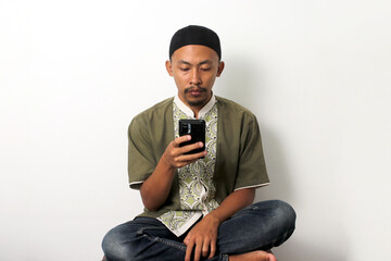 Bored Indonesian Muslim man in koko and peci scrolls through his phone, seeking distraction while...