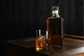 Whiskey with ice cubes in glass and bottle on wooden crate against black background, space for text