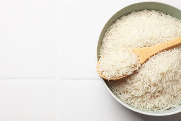 Raw basmati rice in bowl and spoon on white tiled table, top view. Space for text