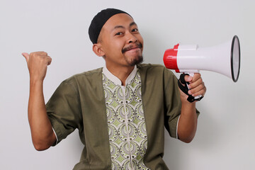 Indonesian Muslim man in koko and peci holds a megaphone and points to the side, directing...
