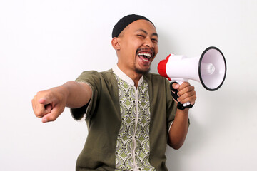 Excited Indonesian Muslim man in koko and peci points at the camera, holding a megaphone to...