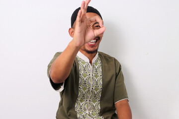 Happy Indonesian Muslim man in koko and peci playfully makes an OK gesture near his eyes, mimicking...