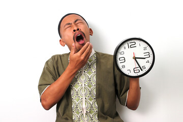 Sleepy Indonesian Muslim man in koko and peci stifles a yawn while checking a clock. He has just...