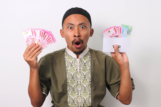 A shocked Indonesian Muslim man with a surprised expression, holds a white envelope labeled THR filled with money, representing his Religious Holiday Allowance. Isolated on a white background