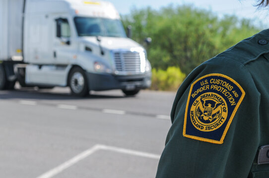 A close-up of the uniform patch of a U.S. Customs and Border Protection agent standing on a road near an interior checkpoint in Southern Arizona, with a passing semi-truck