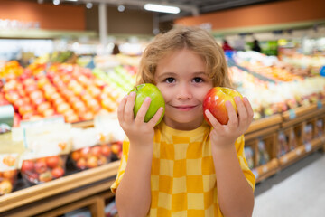 Fototapeta premium Child hold apple fruits at grocery store. Healthy food for kids. Portrait of smiling little child with shopping bag at grocery store or supermarket. Child choosing fruit during shopping at vegetable
