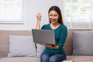 Happy young asian woman relax on comfortable couch at home texting messaging on laptop smartphone, smiling girl use cellphone chatting, browse wireless internet on gadget, work online from home