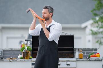Handsome man in shirt and in cook apron preparing fish on barbecue. Male cook cooking salmon fillet...