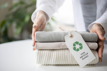 Woman's hands hold a stack of clean neat knitted clothes with a 100% recycle fabric label.