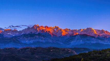 A panoramic view of a mountain range at sunrise, with the peaks bathed in warm yellow sunlight against a backdrop of a deep blue sky