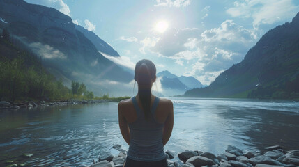 A woman stands in a picturesque outdoor setting surrounded by mountains and a sparkling river. She practices yoga asanas or postures with mindful movements and steady breath