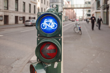 A close-up view of a traffic signal post at an urban intersection, showing a blue bicycle lane light illuminated alongside a red arrow indicating 'no left turn' for vehicles.  - obrazy, fototapety, plakaty