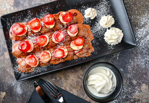 Strawberry and Banana poffers with chocolate, whipped cream, knife and fork served in dish isolated on dark background closeup top view of cafe baked dessert food