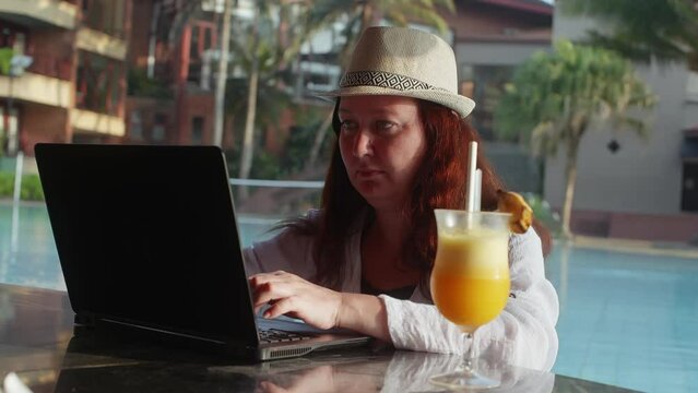 Woman freelancer on vacations in tropics working near pool in hotel drinking cocktail. Middle aged brunette female digital nomad workaholic resting on vacations. Remote work, distant job, freelance.