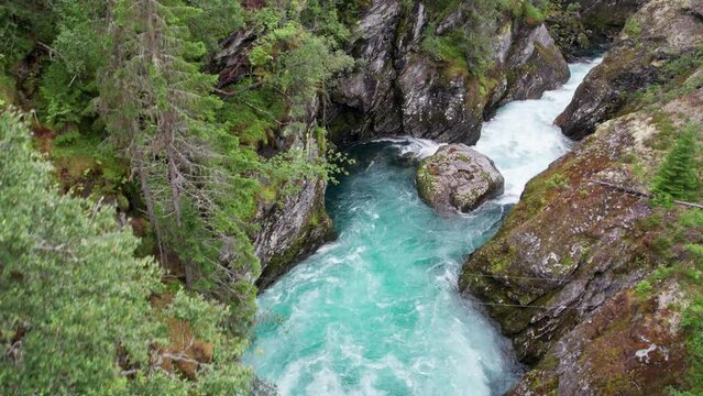 Aerial view, white water rapids rushing between the walls of a craggy, moss covered ravine. Camera climbs and pans, revealing a small road to the left and a pool with still waters in the background