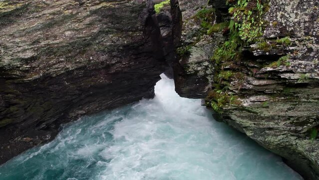 Drone descends into a cave, close to the craggy walls. A powerful waterfall behind a narrow crevice gushes white frothy water into the cave.