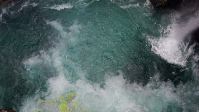 Camera descends vertically into a swirling torrent of white water in a cave, rotates and tilts upwards to reveal a waterfall pouring down through the moss covered rocks of the cave wall.