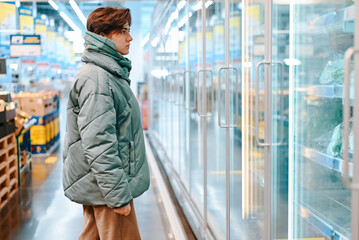 Side view of young woman shopper choosing food in refrigerator in supermarket