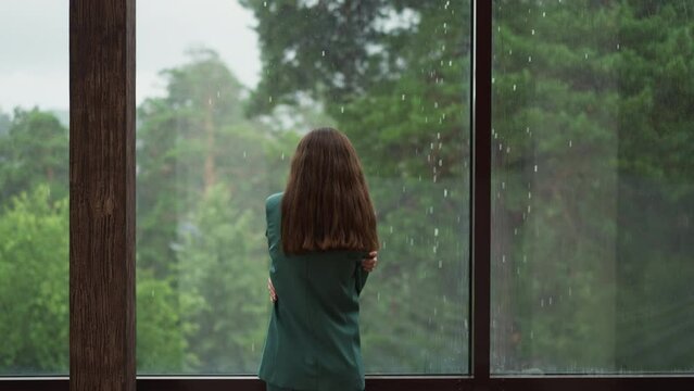 Woman stands in self embrace against window. Woman executive reflects on unrealized potential for company advancement with thoughts clouded by disappointment