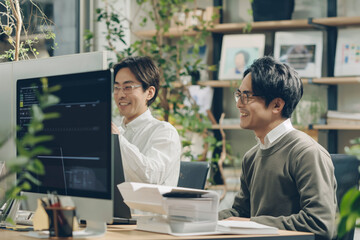Smiling Asian businessman feeling happy at work in office with computer laptop, against office background with plants and partner