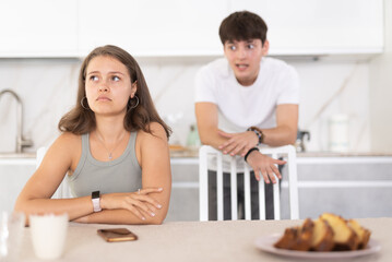 Upset young woman sitting at the kitchen-table with her back to her husband disputing with her