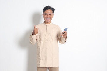 Portrait of young excited Asian muslim man in koko shirt holding mobile phone with smiling...