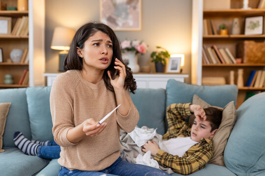 Mother measuring temperature of her ill kid. Sick child with high fever laying in bed and mother holding thermometer. Mother with cell phone calling to doctor