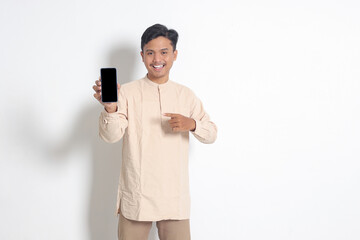 Portrait of young excited Asian muslim man in koko shirt showing blank screen mobile phone mockup while pointing and presenting product. Social media concept. Isolated image on white background