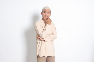 Portrait of confused Asian muslim man in koko shirt with skullcap thinking about question with hand on chin. Education concept. Isolated image on white background