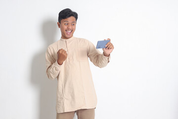 Portrait of excited Asian muslim man in koko shirt holding mobile phone and playing games on his...