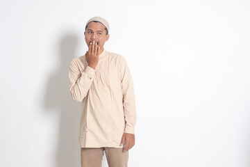 Portrait of overworked Asian muslim man in koko shirt with skullcap yawning with hand covering his mouth. Sleep deprivation concept. Isolated image on white background