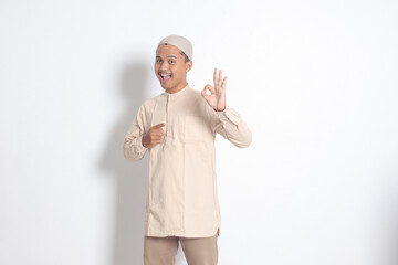 Portrait of young attractive Asian muslim man in koko shirt with skullcap showing ok hand gesture...
