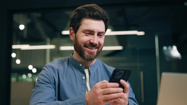 Close up of adult man with beard using modern smartphone and smiling while sitting at coworking space. Satisfied male manager getting great idea while browsing internet and thinking about project.
