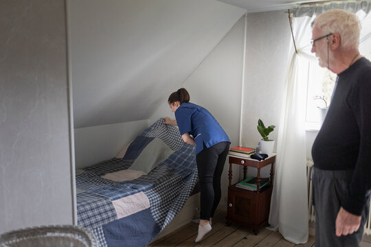 Female nurse spreading sheet on bed for elderly man to sleep in bedroom