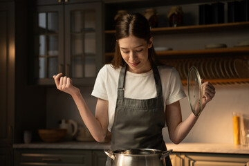 A young woman dressed in an apron stands in a home kitchen, savoring the scent of a dish she is preparing. The gentle evening light filters in through the window, highlighting the steam rising from