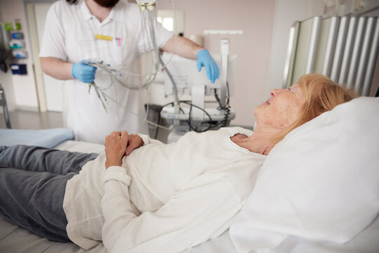 Elderly woman lying on bed by male doctor with electrodes in hospital ward