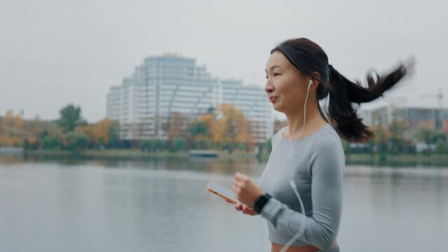 Asian Runner Woman Getting Ready to Run and Starts Running Near the Local Lake, Wearing Earphones and Listening to Music. Woman Training Outside. Jogging And Physical Activity