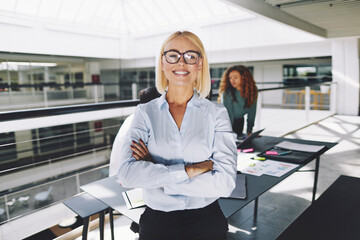 A smiling successful businesswoman looks confidently into the camera while her smiling colleagues are at a meeting in the background